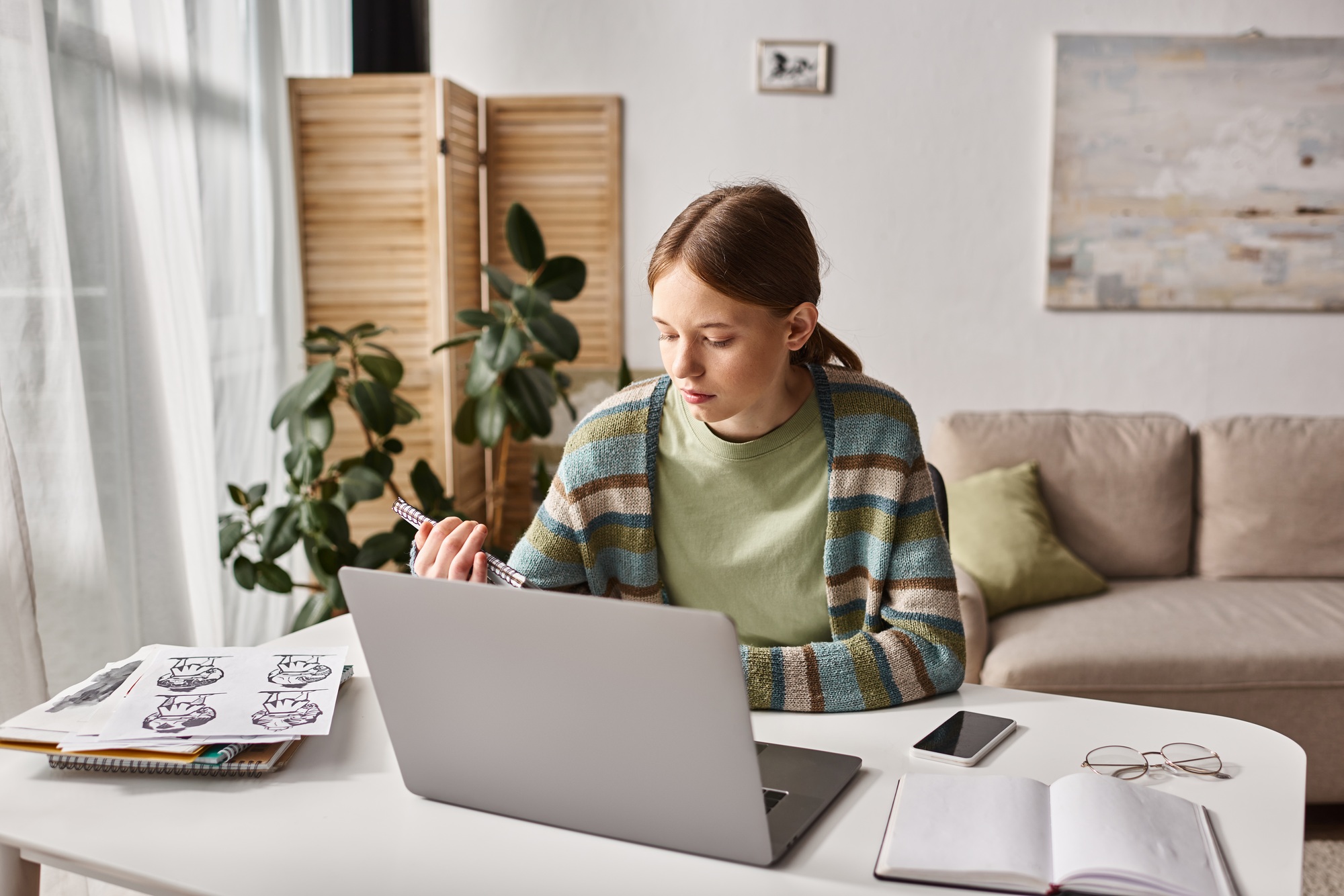 Focused teen girl e-learning with laptop near drawings and smartphone with blank screen on desk