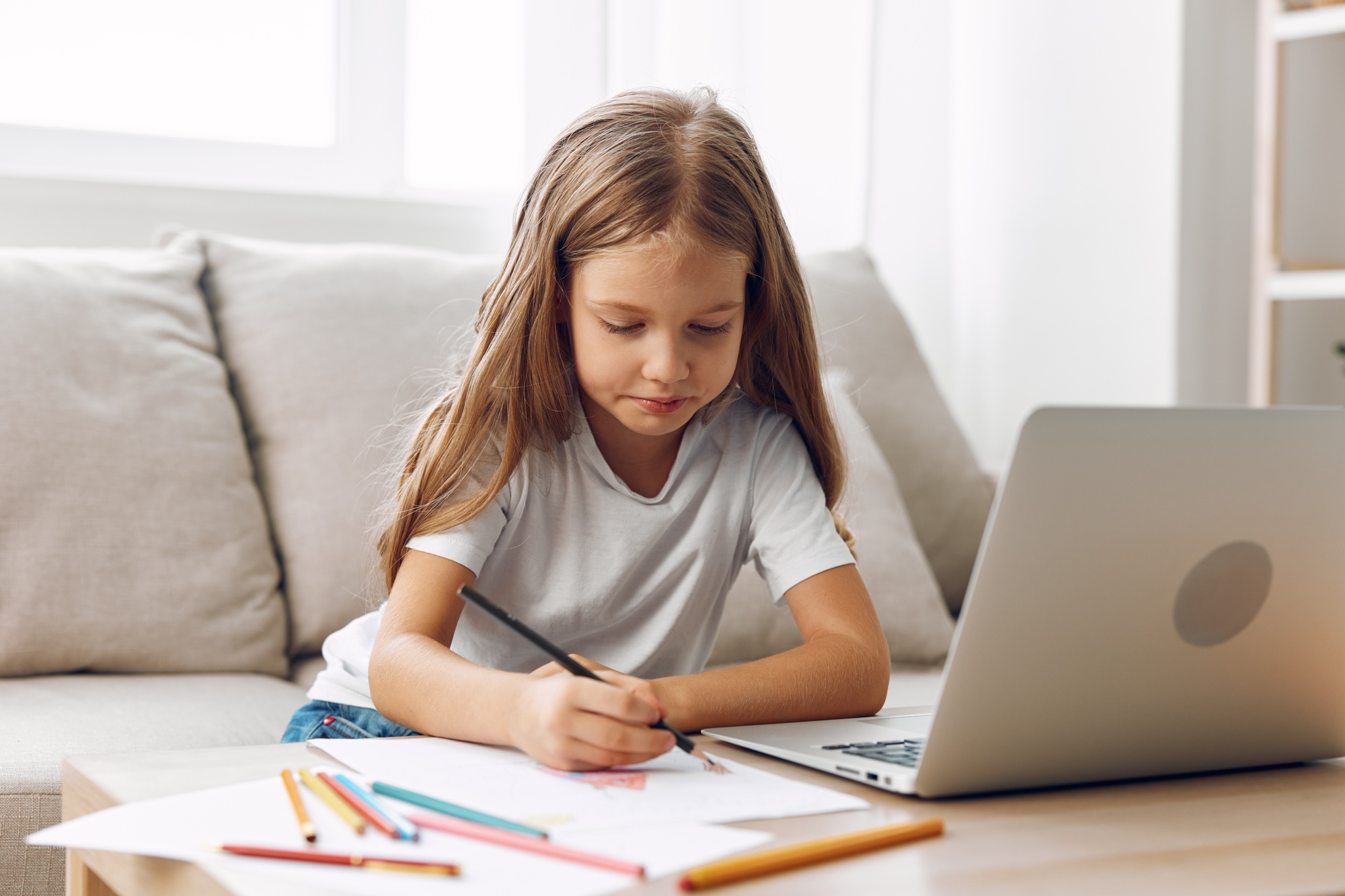 Young girl engaged in creative online learning, sitting at a table with laptop and colorful pencils