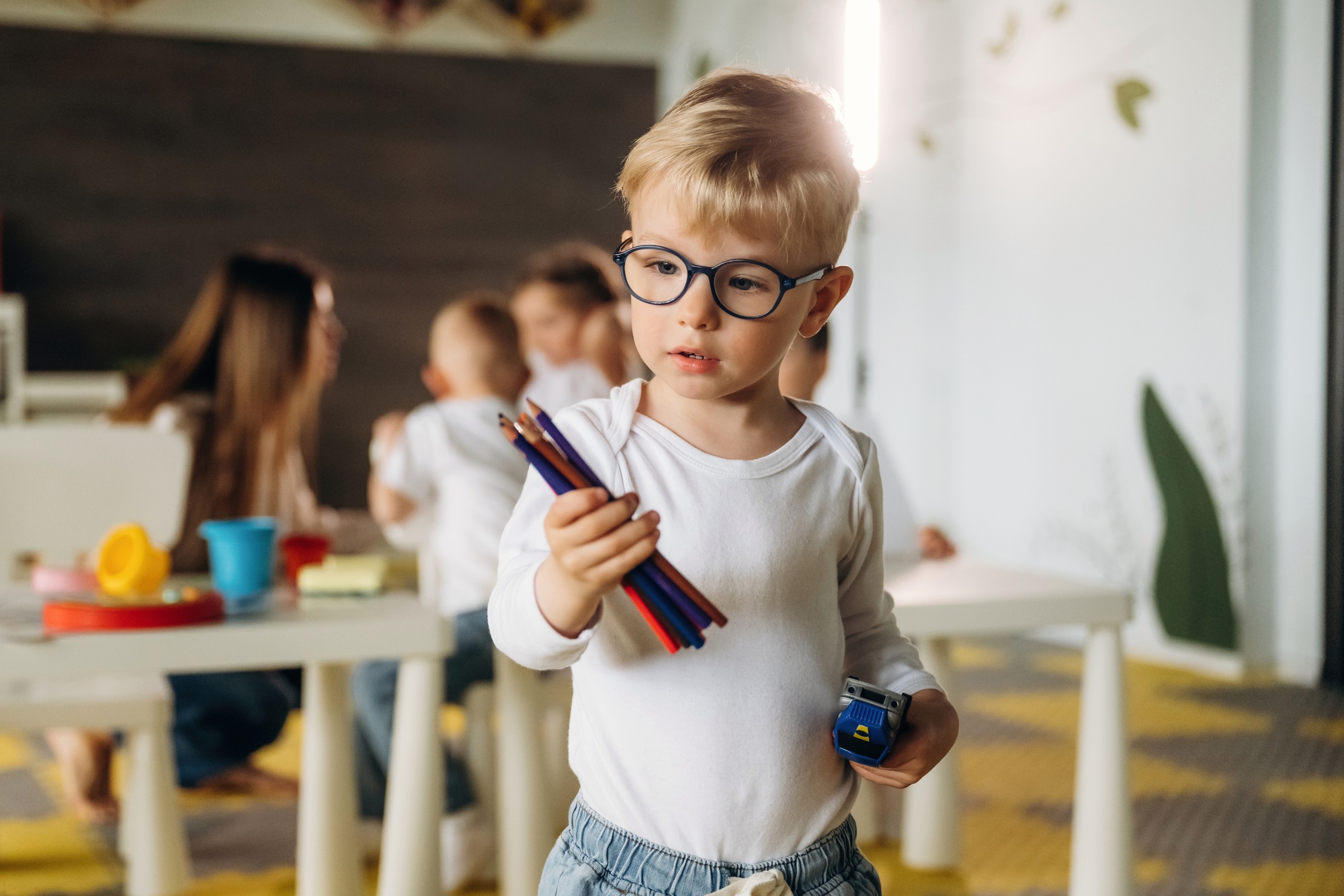 Boy in glasses is standing and holding pencils. Children in kindergarten with their teacher